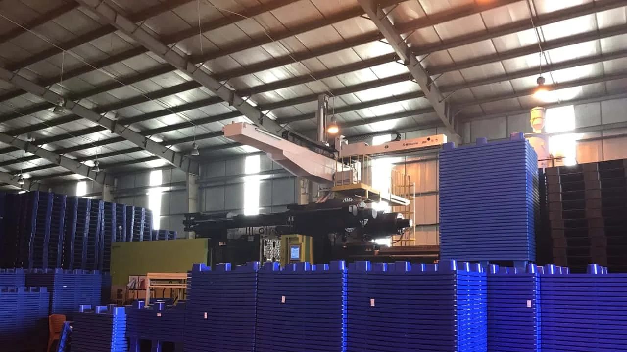 A large injection moulding machine in a workshop, surrounded by stacks of freshly moulded blue plastic pallets