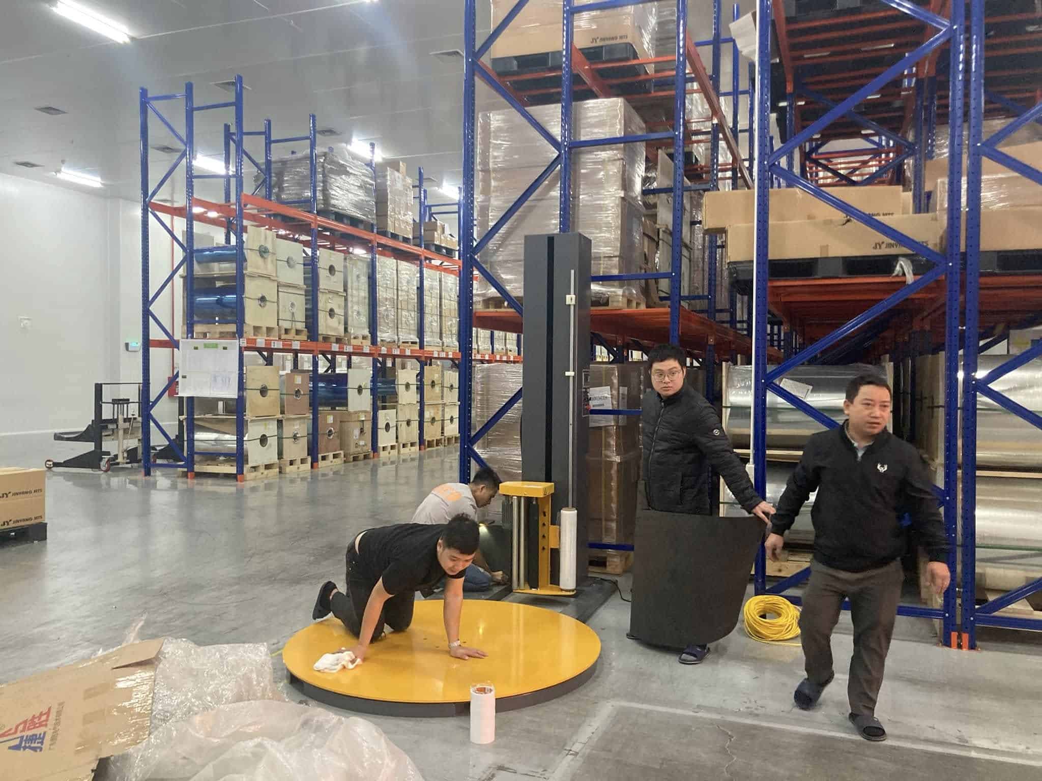 Kỹ thuật viên cân chỉnh mâm xoay màu vàng của máy quấn màng giữa các dãy kệ pallet