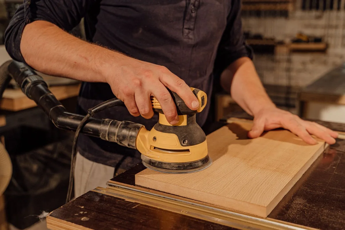 Worker sanding the face of a pale wooden board with an orbital sander
