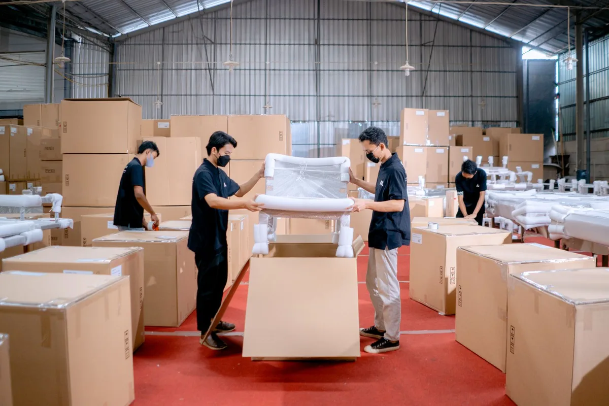 Công nhân đeo khẩu trang đóng ghế bọc màng xốp vào thùng carton trong kho nhà máy nội thất