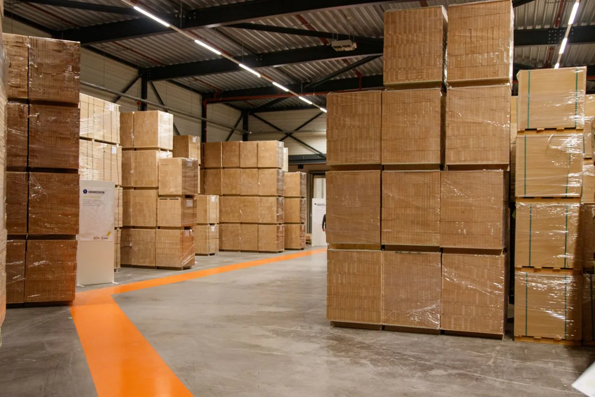 Kho hàng với các khối thùng carton bọc màng xếp trên pallet, nền bê tông có vạch kẻ lối đi màu cam