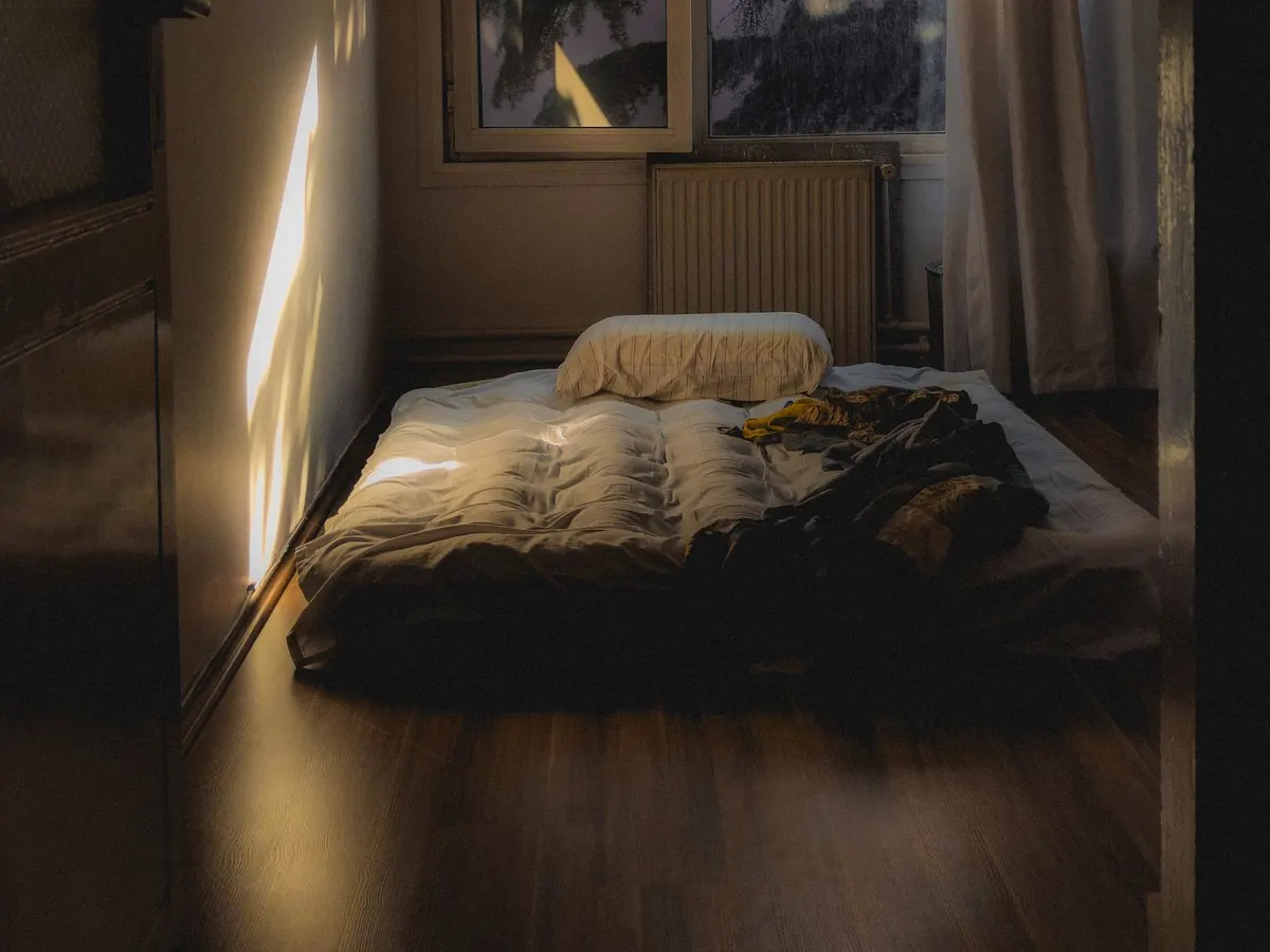 A mattress on the floor in a narrow room with sunlight through the window