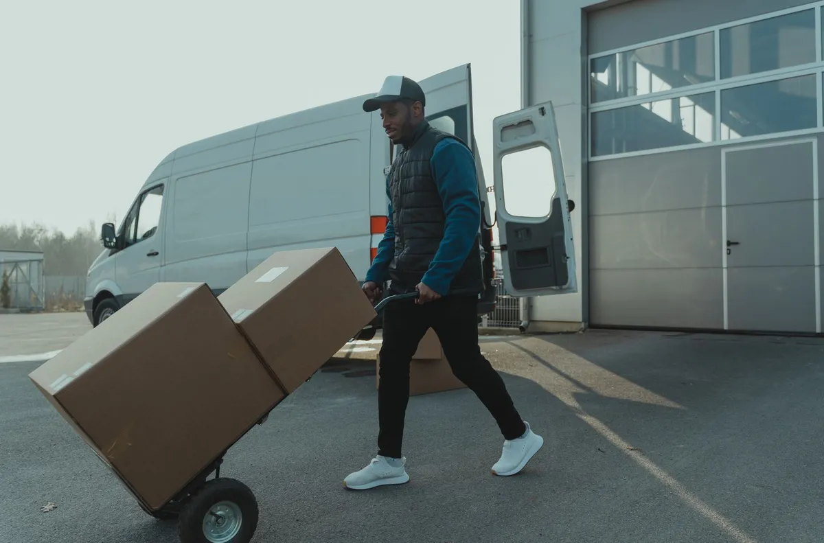 A delivery worker pushing a hand truck carrying two cartons across the yard in front of a warehouse