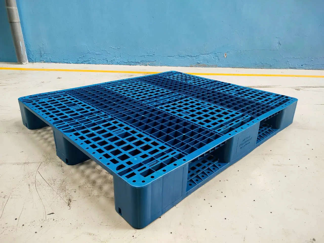 Pallet nhựa 3 chân mặt lưới màu xanh 1300x1100x155 mm đặt trên sàn xưởng, thấy thanh chân chạy dọc thân