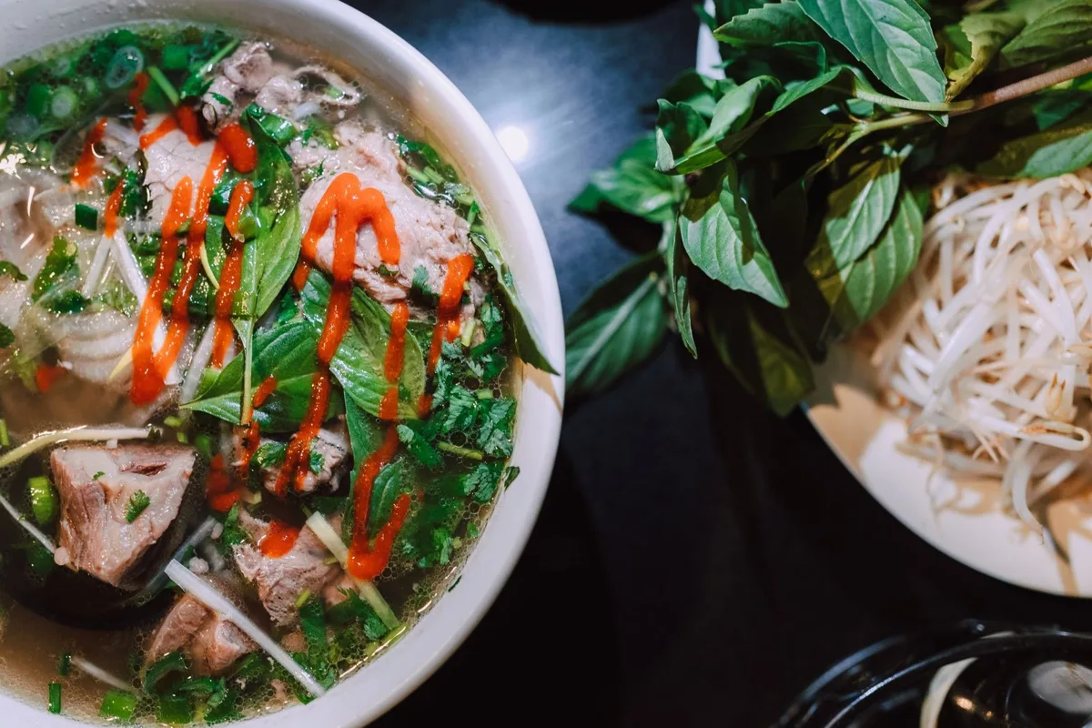 A bowl of beef pho seen from above, next to a plate of herbs and bean sprouts
