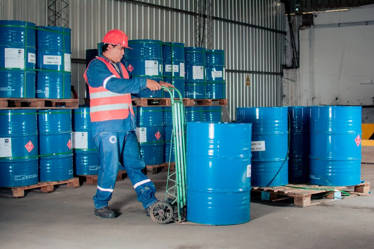 Worker using a trolley to move a blue chemical drum in a warehouse, with drums stacked on pallets