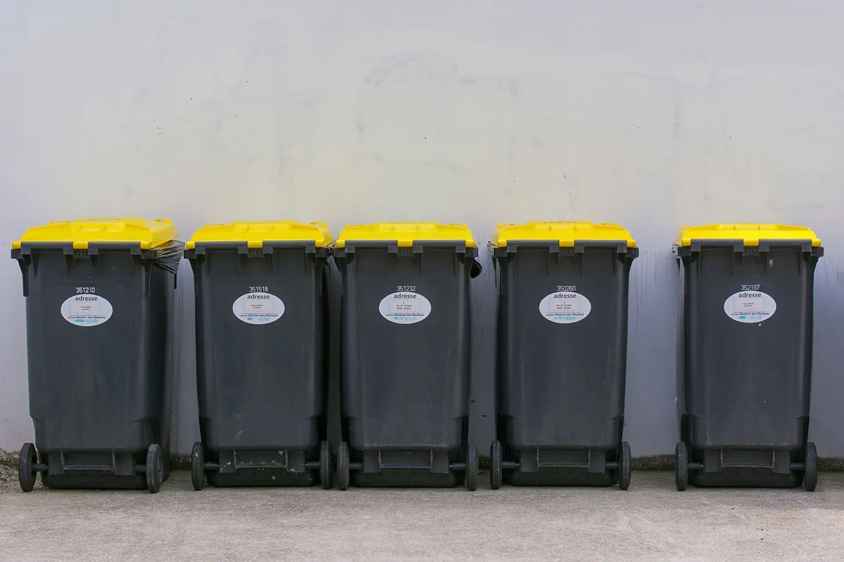 Five grey 2-wheel plastic waste bins with yellow lids lined up against a wall
