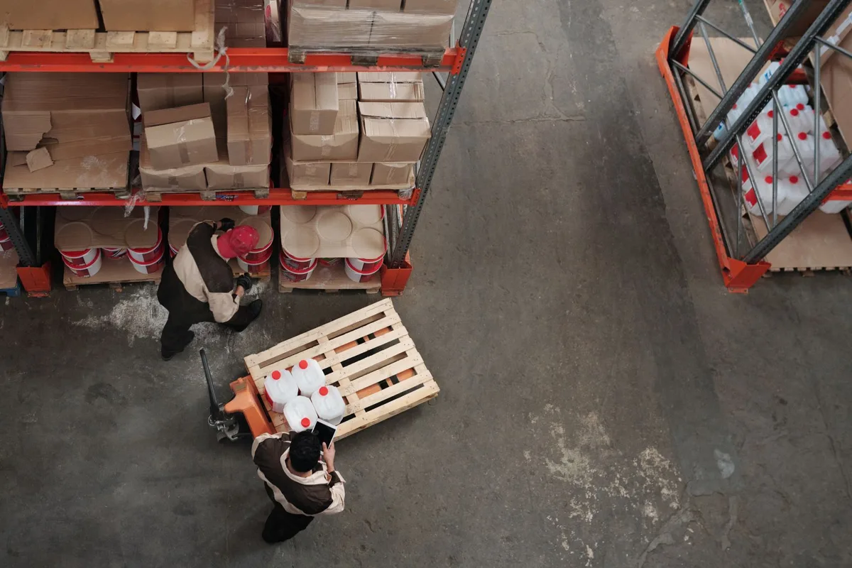 Công nhân kéo pallet hàng bằng xe nâng tay thấp trong lối đi giữa hai dãy kệ